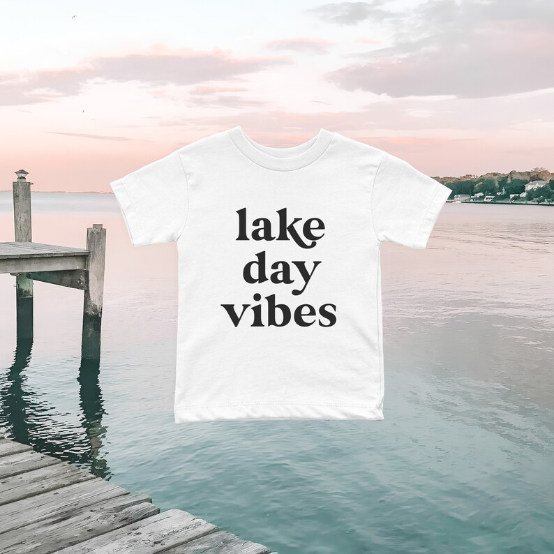 May include: White t-shirt with black text that reads "lake day vibes" against a backdrop of a lake and a wooden dock.