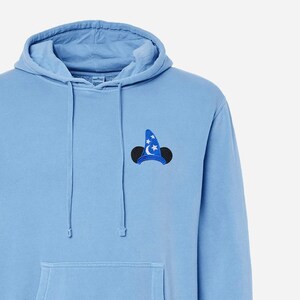 Pu&ograve; includere: Una felpa con cappuccio blu chiaro con una silhouette ricamata di Topolino in nero e blu che indossa un cappello da mago con le stelle.