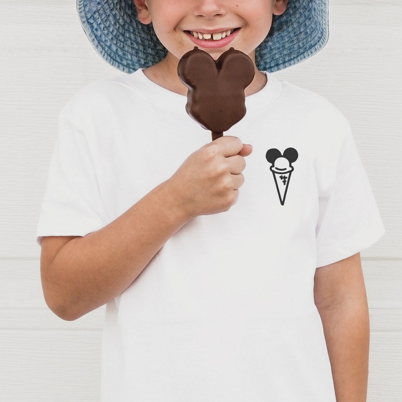 May include: A white t-shirt with a black Mickey Mouse ice cream cone graphic on the front. A child is holding a chocolate Mickey Mouse shaped ice cream on a stick.