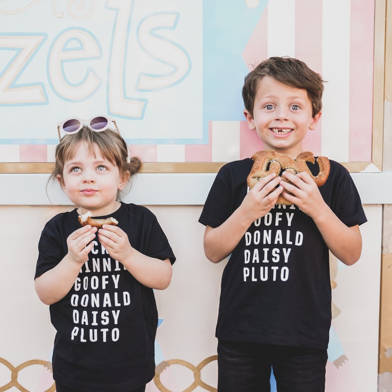 Pu&ograve; includere: Due bambini indossano magliette nere con testo bianco. Il testo sulle magliette recita "Mickey, Minnie, Goofy, Donald, Daisy, Pluto". I bambini tengono in mano dei pretzel e sorridono alla telecamera.