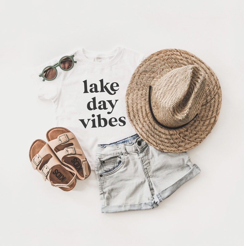 May include: White t-shirt with the text "lake day vibes" in black lettering, a pair of brown sandals, a straw hat, and light blue denim shorts.
