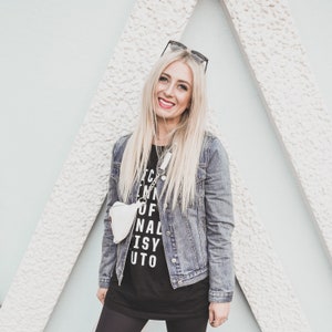 Pu&ograve; includere: Una donna indossa una maglietta nera con la scritta "I'm in of nal isy uto" stampata su di essa, una giacca di jeans e leggings neri. Sorride e indossa occhiali da sole.