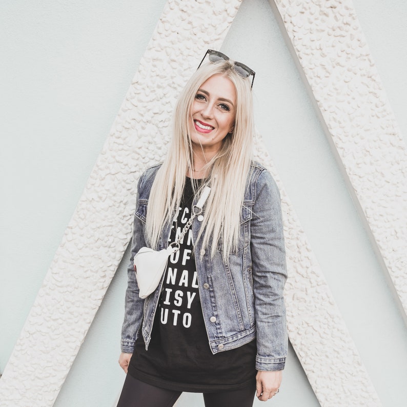 Pu&ograve; includere: Una donna indossa una maglietta nera con la scritta "I'm in of nal isy uto" stampata su di essa, una giacca di jeans e leggings neri. Sorride e indossa occhiali da sole.