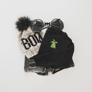 Puede incluir: Una presentaci&oacute;n plana de ropa tem&aacute;tica de Halloween. Incluye un gorro de punto con la palabra "BOO" en negro, una sudadera negra con un gr&aacute;fico verde de Oogie Boogie, pantalones cortos de mezclilla desgastados y una diadema negra con alas de murci&eacute;lago.
