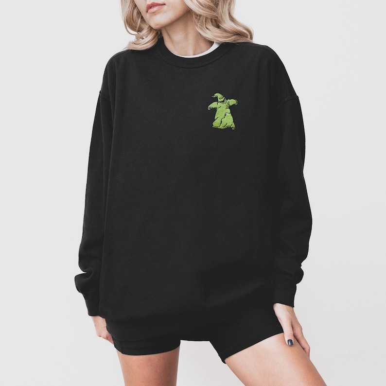 Puede incluir: Sudadera negra con un peque&ntilde;o dise&ntilde;o verde de Oogie Boogie en el pecho. La sudadera tiene cuello redondo y mangas largas. El dise&ntilde;o es un personaje de dibujos animados de la pel&iacute;cula "Pesadilla antes de Navidad".