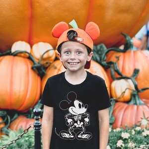May include: A young boy wearing a black t-shirt with a skeleton Mickey Mouse graphic. He is wearing an orange Mickey Mouse hat with large ears. The boy is smiling and standing in front of a pile of pumpkins.