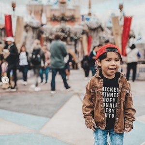 Pu&ograve; includere: Un bambino piccolo che indossa un berretto da baseball rosso, una giacca di jeans marrone, una maglietta nera con la scritta "Mickey Minnie Goofy Donald Daisy Pluto" stampata su di essa e jeans blu. Il bambino &egrave; in piedi davanti a uno sfondo sfocato di un castello.