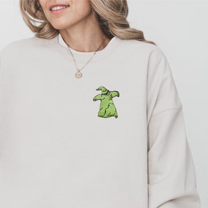 Puede incluir: Sudadera beige con un bordado verde del personaje Oogie Boogie. La sudadera tiene cuello redondo y mangas largas. El dise&ntilde;o de Oogie Boogie est&aacute; centrado en el pecho.