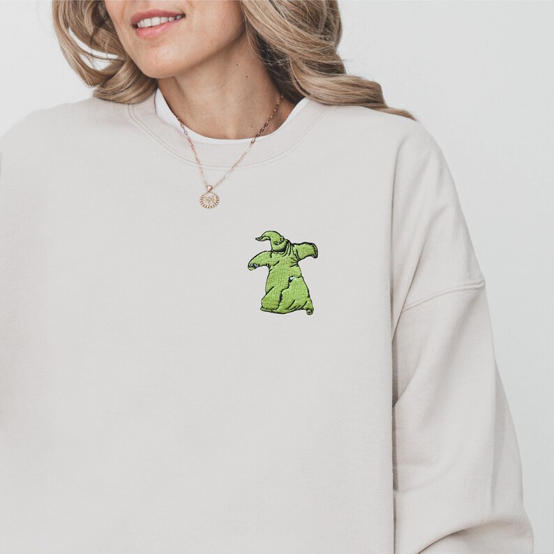 Puede incluir: Sudadera beige con un bordado verde del personaje Oogie Boogie. La sudadera tiene cuello redondo y mangas largas. El dise&ntilde;o de Oogie Boogie est&aacute; centrado en el pecho.
