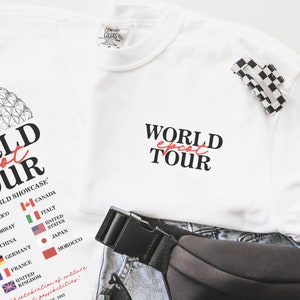 May include: White t-shirt with the text "WORLD EPCOT TOUR" printed in black. The text "EPCOT" is in red. The shirt also features a graphic of the Epcot World Showcase with flags representing different countries.