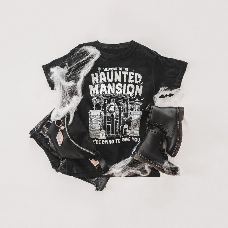 May include: Black t-shirt with a white graphic of a haunted mansion with the text "Welcome to the Haunted Mansion We're Dying to Have You". The shirt is laying on a white surface with a black denim skirt, a black purse with a silver charm, and black boots.