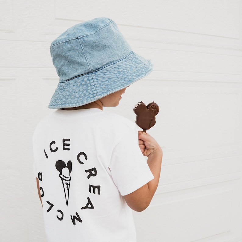 May include: A child wearing a light blue denim bucket hat and a white t-shirt with a black ice cream cone and Mickey Mouse ears graphic that says "Mickey's Ice Cream Club". The child is holding a chocolate Mickey Mouse ice cream bar.