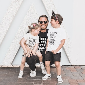 Pu&ograve; includere: Un padre e due figli che indossano magliette abbinate con i nomi dei personaggi Disney stampati su di esse. Le magliette sono bianche con testo nero. Il padre indossa una maglietta nera con lo stesso testo. I bambini sorridono e il padre guarda la fotocamera.