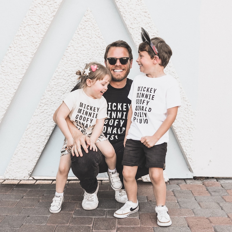 Pu&ograve; includere: Un padre e due figli che indossano magliette abbinate con i nomi dei personaggi Disney stampati su di esse. Le magliette sono bianche con testo nero. Il padre indossa una maglietta nera con lo stesso testo. I bambini sorridono e il padre guarda la fotocamera.