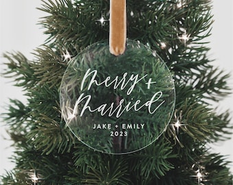 May include: Clear round Christmas ornament with the text "Merry + Married" in white script lettering. Below the text is "Jake + Emily 2023" in black lettering.