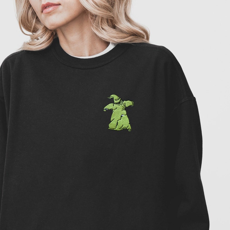 Puede incluir: Sudadera negra con un peque&ntilde;o bordado verde del personaje Oogie Boogie en el pecho. La sudadera tiene cuello redondo y mangas largas. Se ve una camiseta interior blanca en el cuello. El dise&ntilde;o est&aacute; inspirado en la pel&iacute;cula "Pesadilla antes de Navidad".