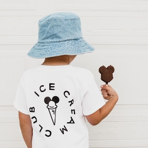 May include: A white t-shirt with a black and white ice cream club logo featuring a cartoon ice cream cone with a Mickey Mouse head. The shirt is being worn by a child wearing a light blue denim bucket hat and holding a chocolate Mickey Mouse shaped popsicle.