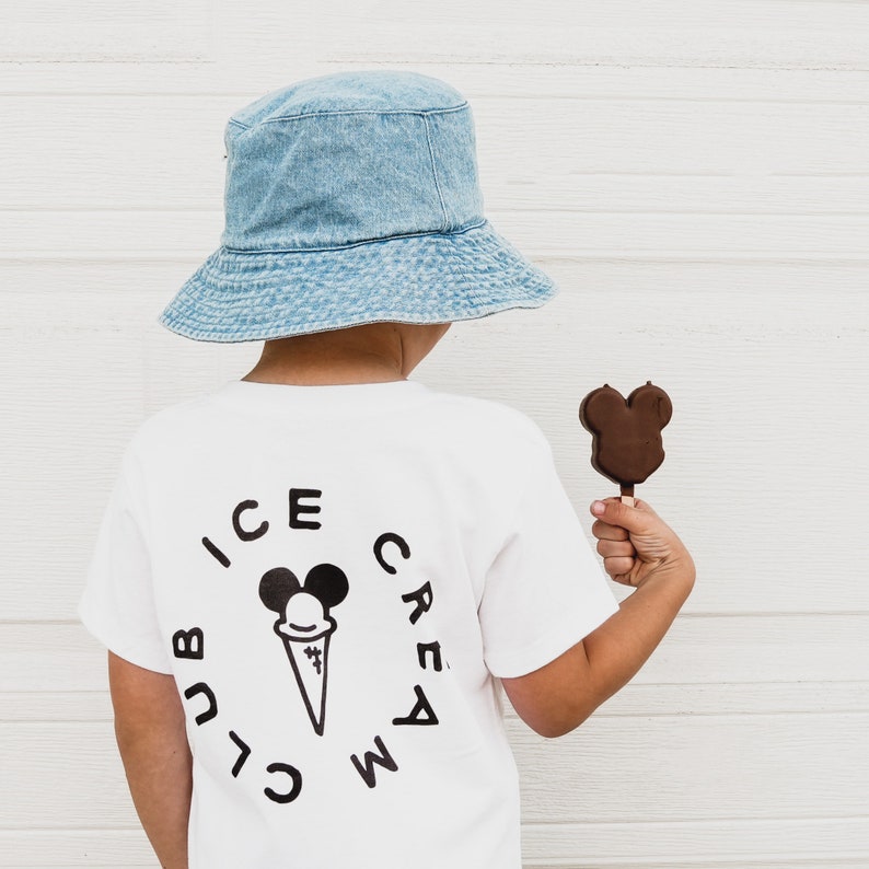 May include: A white t-shirt with a black and white ice cream club logo featuring a cartoon ice cream cone with a Mickey Mouse head. The shirt is being worn by a child wearing a light blue denim bucket hat and holding a chocolate Mickey Mouse shaped popsicle.