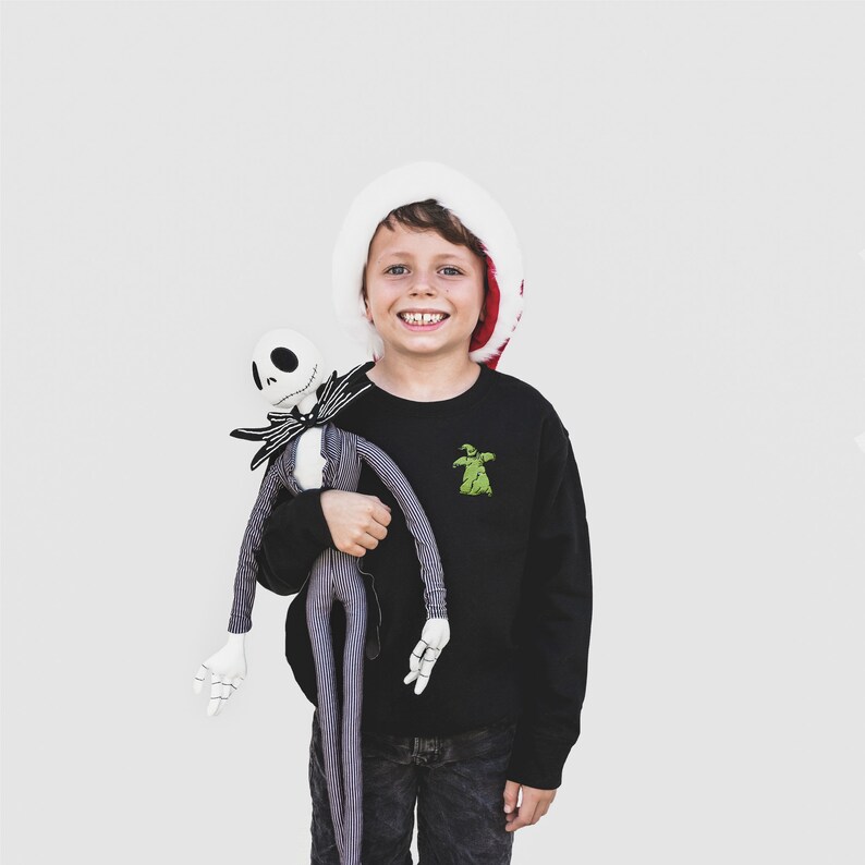 Puede incluir: Un joven lleva una sudadera negra con un gr&aacute;fico verde de Oogie Boogie y un gorro de Pap&aacute; Noel, sosteniendo un mu&ntilde;eco de peluche de Jack Skellington. El mu&ntilde;eco es blanco y negro con un traje a rayas y una pajarita. El fondo es blanco liso.