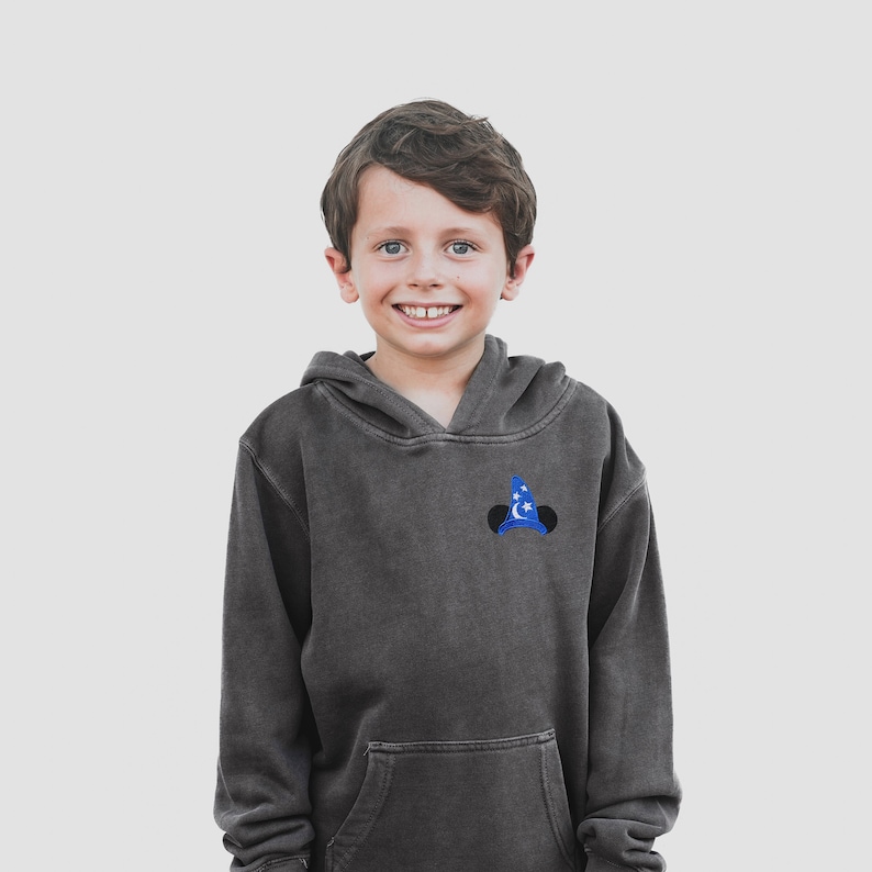 Pu&ograve; includere: Un giovane che indossa una felpa con cappuccio grigia scura con un cappello con orecchie di Topolino ricamato in blu e bianco sul davanti.