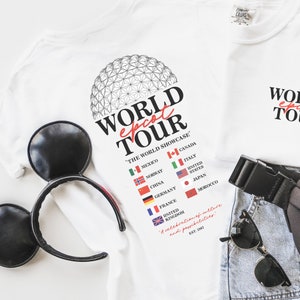 May include: White t-shirt with a black and white illustration of the Epcot World Showcase. The text "WORLD epcot TOUR" is printed in red and black. The text "THE WORLD SHOWCASE" is printed in black. The text "EST. 1982" is printed in black. The t-shirt is a great way to show your love for Epcot.