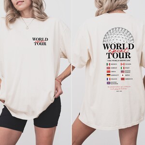 May include: A white t-shirt with a graphic print of a globe and the text "WORLD EPCOT TOUR" on the back. The text "THE WORLD SHOWCASE" is printed below the globe. The globe is surrounded by flags representing different countries, including Mexico, Canada, Norway, Italy, China, United States, Germany, Japan, France, Morocco, and the United Kingdom. The text "A celebration of culture and pavilions" is printed below the flags. The text "EST. 1982" is printed below the celebration text.
