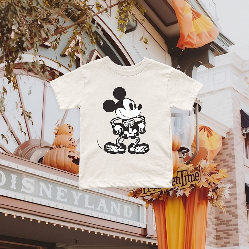 K&ouml;nnte beinhalten: Ein wei&szlig;es T-Shirt mit einem schwarzen und wei&szlig;en Skelett-Mickey-Mouse-Motiv. Das Shirt befindet sich vor einem Geb&auml;ude mit dem Schriftzug "DISNEYLAND".