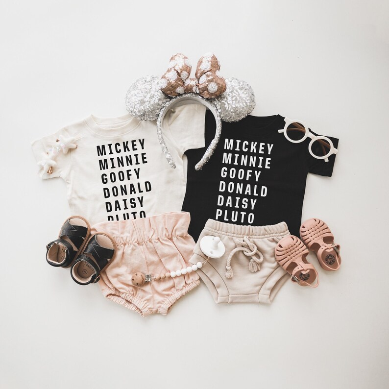 Pu&ograve; includere: Due magliette bianche con testo nero, una maglietta nera con testo bianco, tutte con i nomi dei personaggi Disney: Topolino, Minnie, Paperino, Paperone, Paperina e Pluto. Due paia di bloomers beige per neonati, due paia di sandali marroni, un paio di occhiali da sole bianchi e una fascia rosa e oro con orecchie da topo.