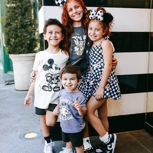 May include: Four children wearing Disney-themed clothing pose in front of a black and white striped wall. The girl in the centre is wearing a black and white checked dress with Mickey Mouse and Minnie Mouse characters. The boy on the left is wearing a white t-shirt with a Mickey Mouse graphic. The boy on the right is wearing a purple t-shirt with a ghost and the text "Spooky Season". The girl on the left is wearing a grey t-shirt with Mickey Mouse characters and a black and white checked headband.