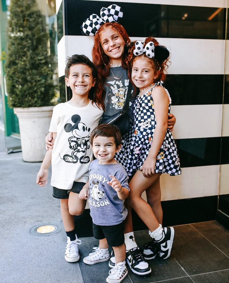 May include: Four children wearing Disney-themed clothing pose in front of a black and white striped wall. The girl in the centre is wearing a black and white checked dress with Mickey Mouse and Minnie Mouse characters. The boy on the left is wearing a white t-shirt with a Mickey Mouse graphic. The boy on the right is wearing a purple t-shirt with a ghost and the text "Spooky Season". The girl on the left is wearing a grey t-shirt with Mickey Mouse characters and a black and white checked headband.