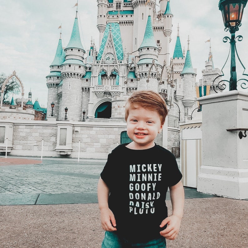 Pu&ograve; includere: Un bambino piccolo che indossa una maglietta nera con i nomi dei personaggi Disney stampati in bianco: Topolino, Minnie, Paperino, Paperone, Paperina, Pluto. Il bambino &egrave; in piedi davanti al Castello di Cenerentola a Walt Disney World.