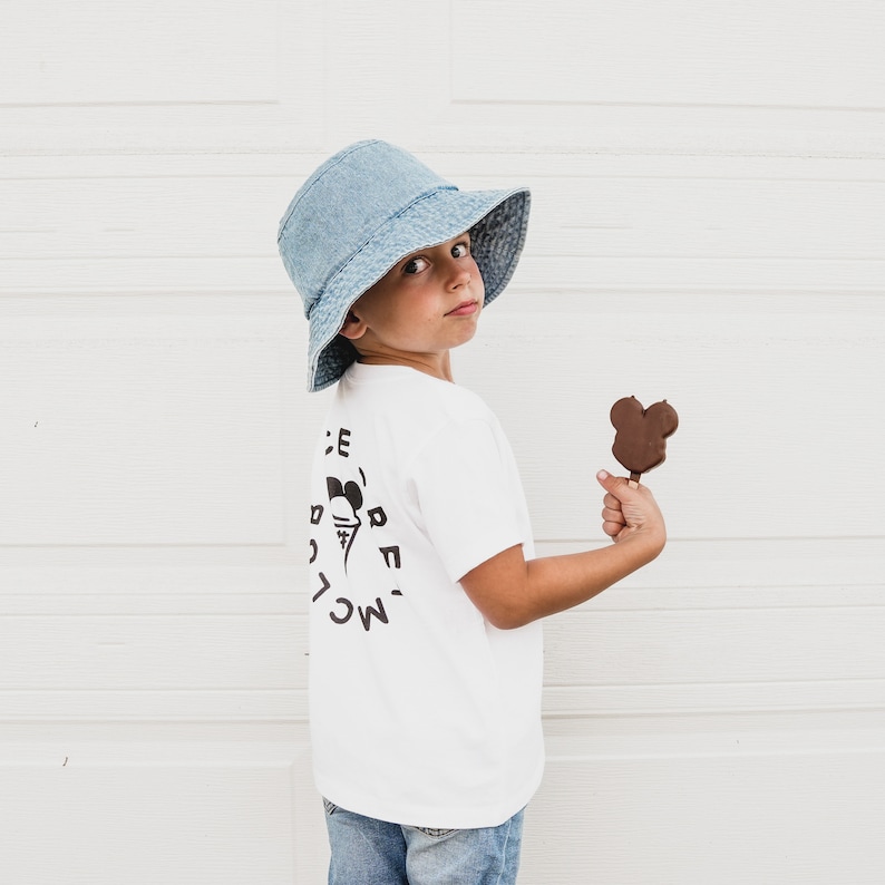 May include: A young person wearing a white t-shirt with a black ice cream cone graphic and the text 'ICE CREAM' on the back. They are wearing a blue denim bucket hat and holding a chocolate Mickey Mouse ice cream bar.