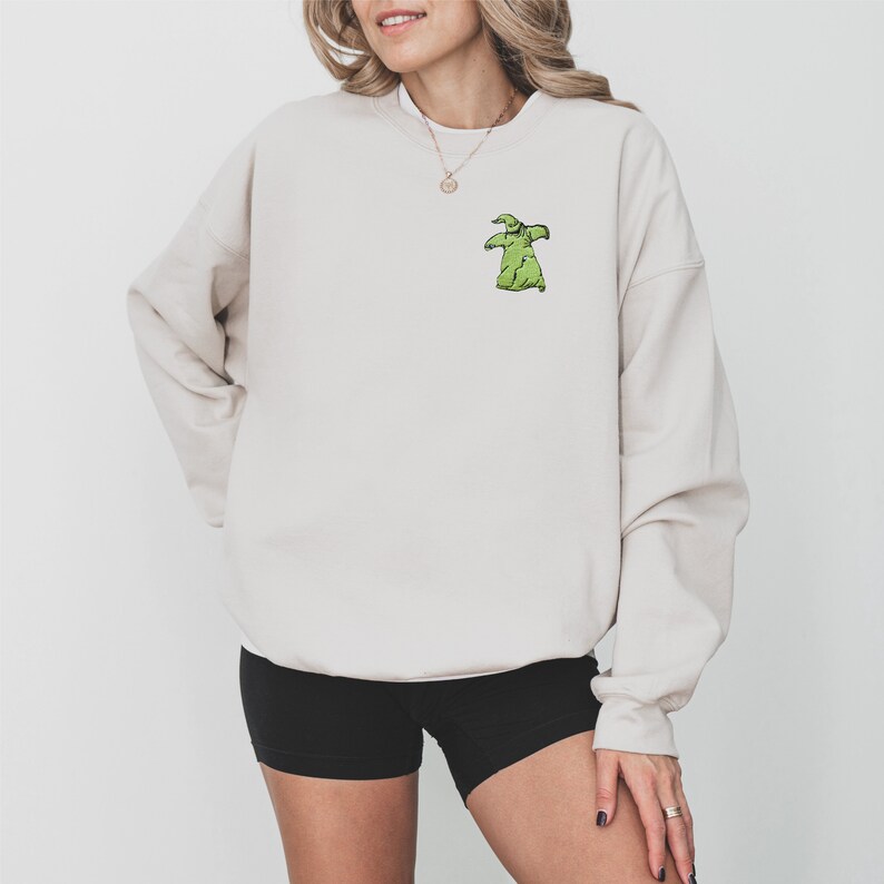 Puede incluir: Sudadera beige con un peque&ntilde;o dise&ntilde;o verde de Oogie Boogie en el pecho izquierdo. La sudadera de manga larga es de color neutro. La persona de la imagen lleva unos pantalones cortos negros.