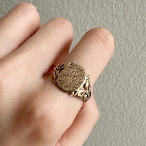 Pode incluir: Um anel de sinete dourado com um monograma gravado. O anel apresenta um design floral ao redor da banda e uma face circular com escrita ornamentada. O anel é usado em um dedo.