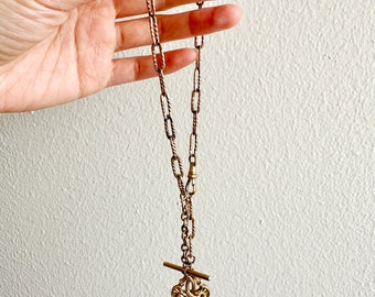 Puede incluir: Collar antiguo de color dorado con un colgante decorativo. El collar presenta una cadena larga con una barra en T y un colgante ornamentado. La cadena está hecha de eslabones entrelazados, el colgante tiene un diseño detallado.