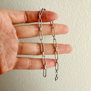 Puede incluir: Una pulsera de cadena plateada con eslabones rectangulares alargados. La pulsera está sujeta por una mano sobre un fondo blanco. La pulsera tiene un cierre de mosquetón.