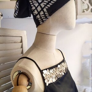 May include: A black sleeveless top with a decorative neckline made of white shell-like embellishments. The top is worn on a mannequin with a black and white patterned head scarf.
