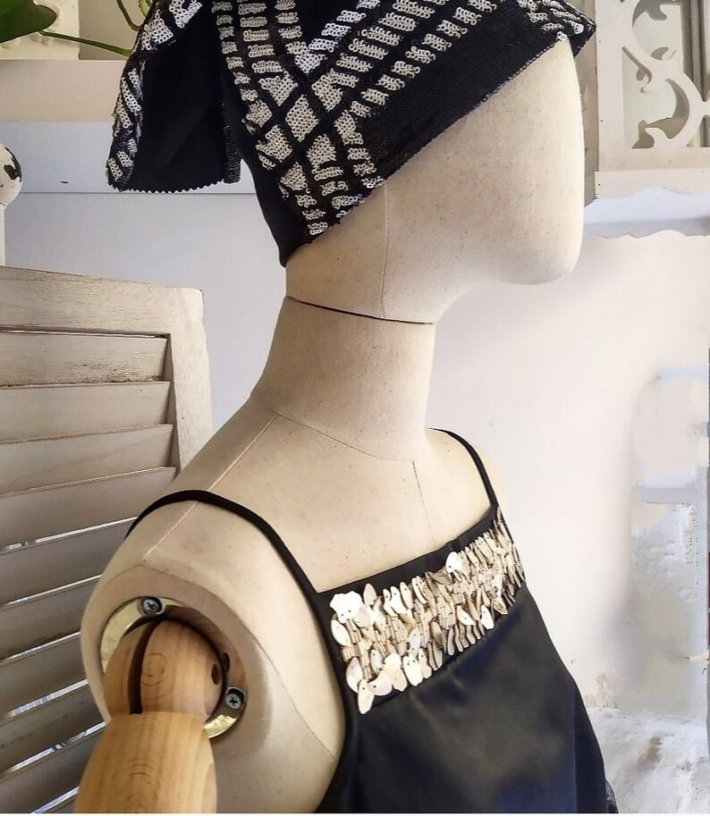 May include: A black sleeveless top with a decorative neckline made of white shell-like embellishments. The top is worn on a mannequin with a black and white patterned head scarf.