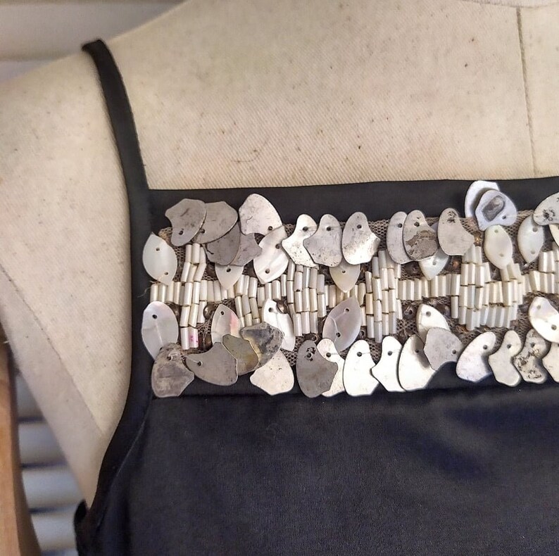 May include: A black sleeveless top with a silver and white beaded trim. The trim features a row of small, silver, teardrop-shaped sequins and a row of white beads.