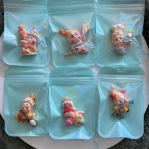 May include: Six colorful, cartoonish figurines in individual clear plastic bags. Each figurine depicts a character in various poses, wearing hats and costumes. The figurines are in shades of yellow, orange, pink, and blue.
