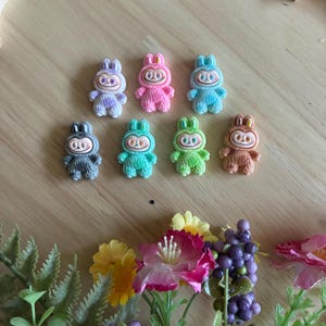 May include: Seven small, textured, rabbit-shaped figurines in various colors including lavender, pink, light blue, gray, teal, lime green, and tan. Each figurine features a smiling face with large eyes and upright ears, arranged in two rows.