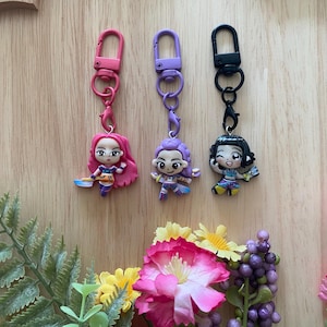 May include: Three colorful keychains featuring miniature cartoon figures with pink, purple, and black hair. Each keychain has a matching carabiner clip. The figures are holding various accessories and are attached to a wooden surface.