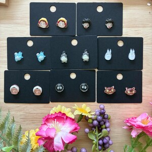 K&ouml;nnte beinhalten: Eine Sammlung von Ohrsteckern mit Charakteren aus einem beliebten Zeichentrickfilm. Die Ohrringe werden auf schwarzen Karten pr&auml;sentiert, mit Designs, die Charaktere mit gelben Haaren, schwarzen Haaren und katzenartigen Gesichtern zeigen. Die Ohrringe sind farbenfroh und detailliert.