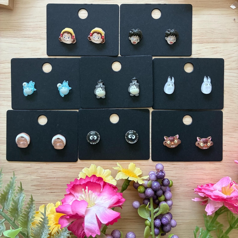 K&ouml;nnte beinhalten: Eine Sammlung von Ohrsteckern mit Charakteren aus einem beliebten Zeichentrickfilm. Die Ohrringe werden auf schwarzen Karten pr&auml;sentiert, mit Designs, die Charaktere mit gelben Haaren, schwarzen Haaren und katzenartigen Gesichtern zeigen. Die Ohrringe sind farbenfroh und detailliert.