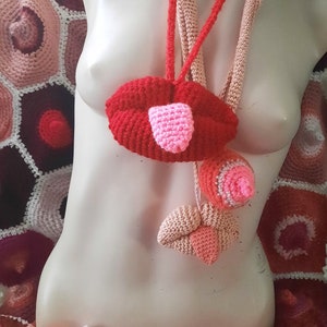 May include: A crocheted necklace with a red lip, pink tongue, and two pink and white crocheted balls hanging from it.
