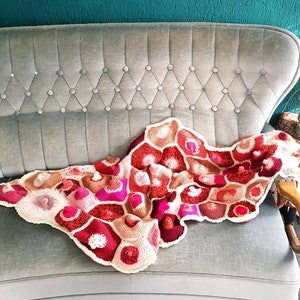 Könnte beinhalten: Eine gehäkelte Wolldecke mit einem geometrischen Muster in Rosa-, Rot- und Beigetönen. Die Decke liegt auf einem grauen Sofa.