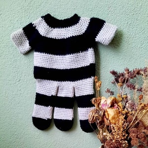 May include: A black and gray striped crocheted baby outfit with long sleeves and a mitten-like design on the hands. The outfit is hanging on a light green wall.