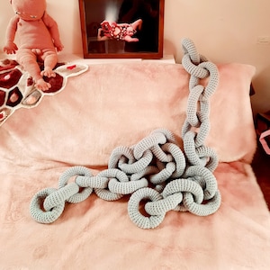 May include: A light blue crocheted chain links on a pink fluffy surface. The chain links are arranged in a loose, organic pattern.