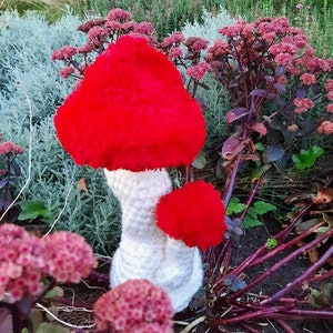 Könnte beinhalten: Zwei gehäkelte Pilze mit roten Hüten und weißen Stielen. Die Pilze stehen in einem Gartenbeet mit rosa Blüten.