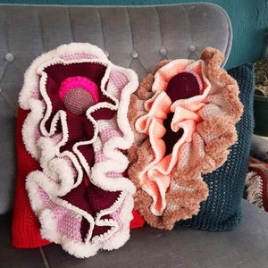 May include: Two crocheted pillows with ruffled edges. One pillow is pink, white, and burgundy with a small pink and white crocheted ball in the center. The other pillow is peach, white, and brown with a burgundy crocheted ball in the center. Both pillows are on a gray couch.